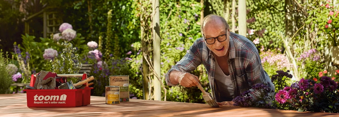 Eine Person streicht mit einem Pinsel Holzdielen in einem blühenden Garten.